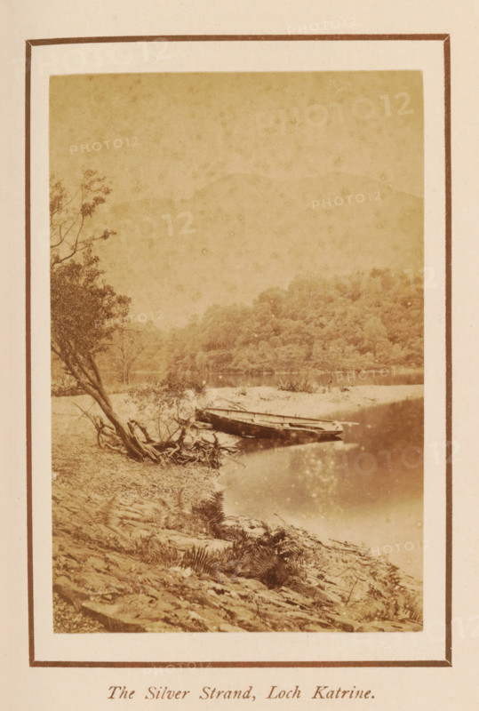 The Silver Strand, Loch Katrine, photograph, page, photographer: George Washington Wilson, Scotland, c. 1885 - c. 1890, paper, albumen print, height 104 mm x width 69 mm, lake, ships, boats on land, beached, A sepia-toned photograph bordered by a double mat shows a calm shoreline scene with a gently sloping rocky bank in the foreground, a fallen tree with exposed roots leaning toward the water on the left, and a small wooden boat pulled up at the water's edge. The water reflects light and the opposite shore, where a densely wooded hillside rises, and faint tree branches frame the left side of the image. The overall image has a textured, aged appearance.