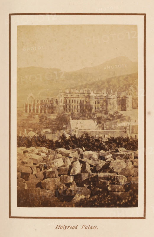 Holyrood Palace, photograph, page, photographer: George Washington Wilson, Edinburgh, c. 1885 - c. 1890, paper, albumen print, height 102 mm x width 69 mm, palace, façade (of house or building), Palace of Holyroodhouse, A sepia-toned photograph framed by a pale border shows a distant large stone building with multiple windows and towers set against rolling hills, with a ruined arched structure visible to the left of the main building; in front of the building there are smaller roofs and tree foliage, and the foreground is filled with a jumble of irregular stones and rocks.