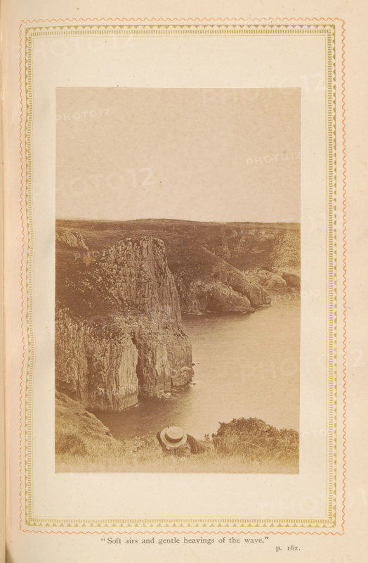 Landscape with rock wall and a man, photograph, page, inscription: ''Soft airs and gentle heavings of the wave.', p. 162.', c. 1885 - in or before c. 1890, photographer, paper, albumen print, albumendruk, height 108 mm x width 69 mm, coastal cliff or crag (landscape with figures, staffage), A sepia-toned photograph bordered by decorative patterned margins shows a rugged coastal cliff dropping steeply to a calm sea, with layered rock faces and grassy terrain at the top. The coastline curves into a sheltered inlet where rocky outcrops meet the water. In the foreground, a person wearing a wide-brimmed straw hat reclines on the grassy slope facing the sea, with nearby low shrubs and grasses around them. The sky above the distant cliffs is pale and expansive.