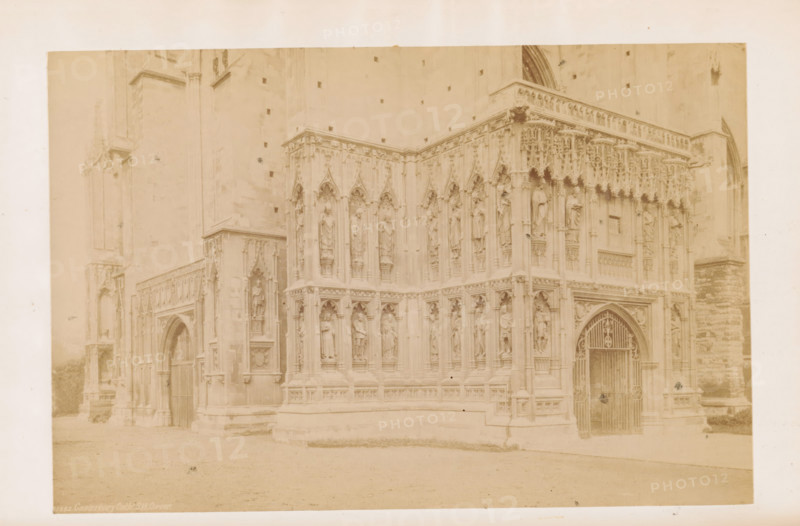 The Cathedral, Southwest Corner, View of Canterbury Cathedral, photograph, page, Canterbury Cathedral, c. 1882 - in or before 1892, photographer, Kent, Canterbury, paper, albumen print, height 115 mm x width 174 mm, church (exterior), parts of church exterior and annexes: portal, A faded sepia photograph showing a stone building corner with ornate Gothic carvings and sculpted figures set in niches across two levels, pointed arches, and decorative tracery; a gated arched doorway with an iron gate is visible on the right side, flanked by carved columns and statues; the left side shows another arched entrance partially recessed beneath detailed stonework; the ground in front appears smooth and level, and the upper facade shows small square openings and a plain stone wall above the carved section.