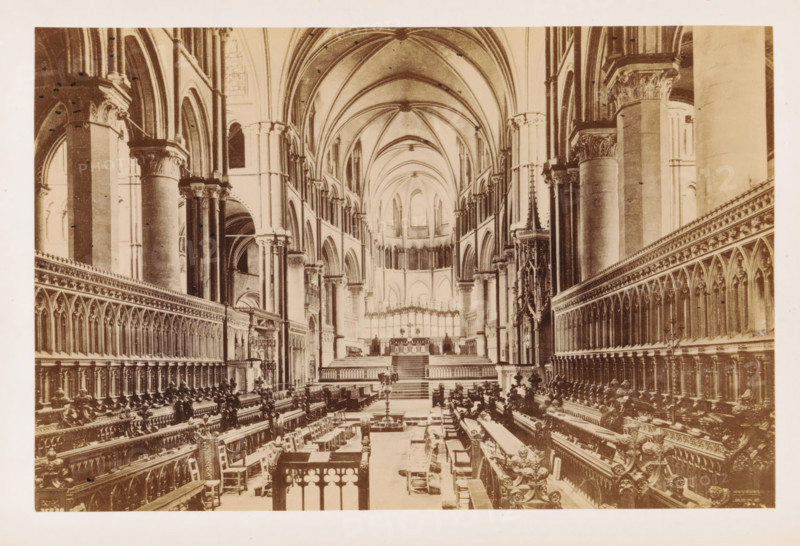 The East Choir, Interior of Canterbury Cathedral with a view of the choir, photograph, page, Canterbury Cathedral, c. 1882 - in or before 1892, photographer, Kent, Canterbury, paper, albumen print, height 115 mm x width 175 mm, parts of church interior: choir, A wide interior view of a large cathedral nave with tall vaulted ceilings and pointed arches, flanked by rows of ornate wooden choir stalls with carved details and high backs on both sides, a central aisle leading toward an elevated chancel area with steps and an ornate screen, massive stone columns with decorated capitals supporting the arches, slender clerestory windows high above, decorative stone tracery and carved ornament along the walls, scattered chairs and a few candlesticks or stands along the aisle, and architectural details extending symmetrically toward a distant apse.
