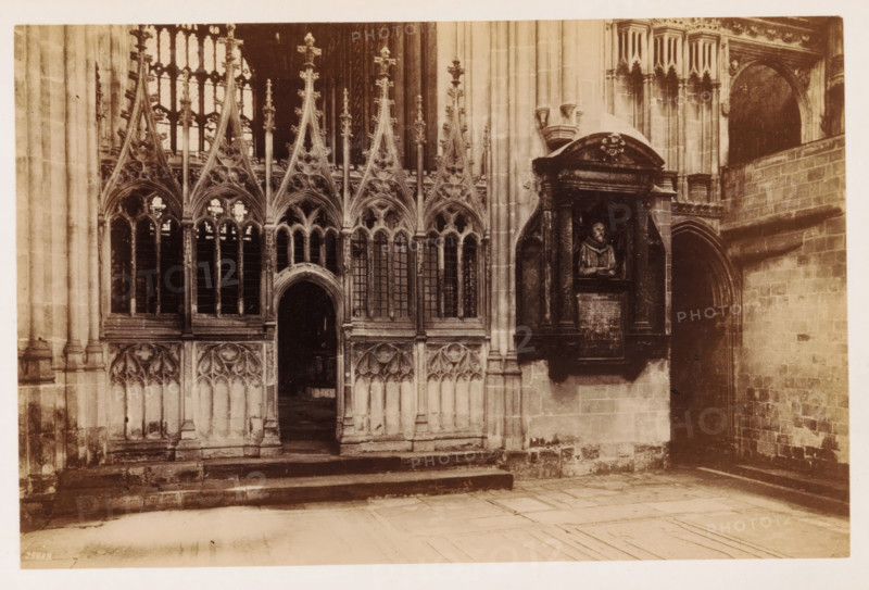 Interior of a transept of Canterbury Cathedral, The Transept of the Martyrdom, photograph, page, number: '25689', Canterbury Cathedral, c. 1882 - in or before 1892, photographer, Kent, Canterbury, paper, albumen print, height 115 mm x width 175 mm, parts of church interior: transept, A sepia-toned photograph of an ornate stone interior showing a series of pointed Gothic arches with tracery and tall narrow windows, ornate finials and carved vertical ribs, and a central arched doorway leading into a darker interior. To the right of the arches a sculpted wall monument sits within a recessed niche framed by columns and an arched pediment, featuring a carved bust set above a rectangular plaque. The surrounding walls are composed of dressed stone blocks, with a secondary arched passageway visible on the far right and worn stone steps and a tiled floor in the foreground.
