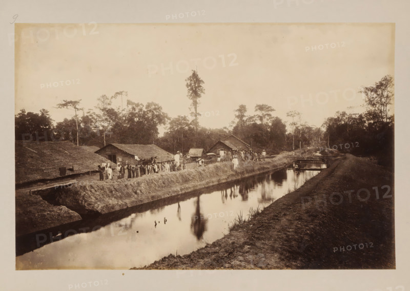 Canal at the Stabat plantation, Sumatra, album page, photograph, This photo is part of an album., number: '9', photographer, c. 1890 - c. 1900, paper, pasteboard, albumen print, height 243 mm x width 372 mm, In the list of images, this photo is described with the words 'Kanal in Stabat Estate'., canal, plantation, working class, labourers, A sepia-toned photograph showing a narrow waterway reflecting trees and the sky, with earthen banks on both sides and a dirt path along the near bank, a group of people standing along the far bank near thatched-roof huts and simple wooden structures, a few carts and a person seated on one of them, clusters of trees rising behind the huts including a tall solitary tree, a small wooden footbridge crossing the water farther down, and a person seated on the bridge while another stands nearby.