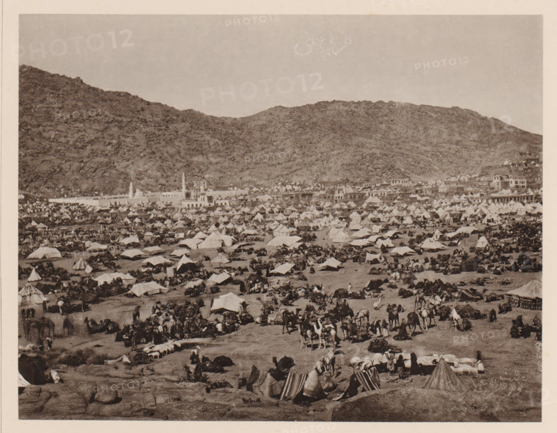 Pilgrim camp in Mina near Mecca during the Hajj, Westlicher Theil des sich von Westen nach Osten erstreckenden Thales Muna (Mina) während der grossen jährlichen Pilgerversammlung, album page, photomechanical print, This print is part of an album., number: 'X', photographer: Abd al-Ghaffar, Mina, cliché maker: anonymous, 1889, Saudi Arabia, pasteboard, paper, collotype, photoprint, height 270 mm x width 360 mm, tent-camp, Hajj, Period, 1888-03-30 - 1888-04-27, A wide landscape shows a crowded encampment spread across a flat area at the base of rocky hills, with numerous tents of various shapes and sizes clustered closely together and rows of low buildings along the edge of the settlement; many people are scattered among the tents, some standing, some seated, and groups are gathered around camels and packs, while camels lie or stand throughout the foreground; the scene is monochrome and framed by a pale border, with rugged hills rising behind the settlement and a clear sky above.