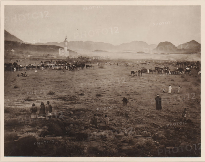 Die zwischen Muna und Arafah situated Pilgerstation Muzdal'fah, View of Muzdalifah, located between Mina and the plain of Arafat, during the Hajj, album page, photomechanical print, This print is part of an album., number: 'XII', photographer: Abd al-Ghaffar, Muzdalifah, cliché maker: anonymous, 1889, Abd al-Ghaffar, photographer, cliché maker, Saudi Arabia, pasteboard, paper, collotype, photoprint, height 270 mm x width 360 mm, other ritual practices, Islam, Mohammedanism, Hajj, Period, 1888-03-30 - 1888-04-27, A wide, sepia-toned landscape shows a flat, rocky plain stretching toward distant hazy mountains under a pale sky, with a tall tower and adjacent building near the middle left skyline. Groups of people are scattered across the plain, some standing, some sitting, and several small clusters of animals, including pack animals, are gathered near the center and right. In the foreground three figures stand together facing away, and a few other solitary figures sit or stand on rocky g...