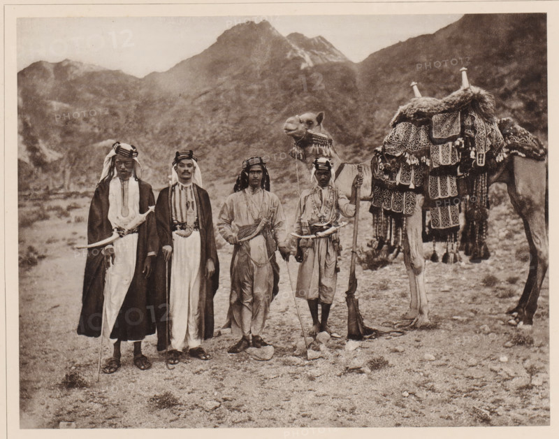 Das Reitkamel (Hèdjin) des Scherif Jahja, eines Sohnes des Scherif Ahmed, dessen Vater der beruhmte 1886 verstorbene Grossscherif Abd èl-Muttálib with a grain of silver gestickten Satteldecke (batat), the Zaum hält der Sklave Jahja's, neben diesem steht Jahja selbst im Reiteranzug and on the left in long Kleideren zwei Scherife niedrigeren Ranges, Group portrait of sjarif Jahja with on the right an enslaved boy with a dromedary and on the left two unknown sjariffs, album page, photomechanical print, This print is part of an album., number: 'XVII', photographer: Abd al-Ghaffar, Saudi Arabia, cliché maker: anonymous, 1889, Abd al-Ghaffar, photographer, cliché maker, pasteboard, paper, collotype, photoprint, height 270 mm x width 360 mm, Person, Jahja, historical persons, historical persons not known by name, hoofed animals: dromedary, family of the sovereign, Four men stand side by side in traditional robes and head coverings, each holding a staff or weapon, with one man wearing a long d...