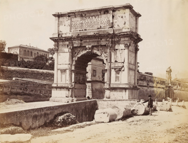 Man posing by the Arch of Titus at the Roman Forum in Rome, photograph, Part of a French artist's album., Rome, c. 1880 - c. 1900, photographer, paper, albumen print, height 194 mm x width 254 mm, triumphal arch, Roman Forum, Arch of Titus, A large stone triumphal arch with a central arched opening stands prominently in the scene, showing carved reliefs, fluted columns, and weathered surfaces with visible cracks and dark stains; fragments of broken columns and cylindrical stone drums lie on the ground in front of the arch; a man wearing a dark coat and hat stands beside the stone fragments; a low stone wall and walkway run alongside the arch and foreground; behind and to the left of the arch there is a multi-windowed building partially obscured by trees and shrubbery; to the right a long wall with decorative statues and architectural details extends into the distance; the overall image has a sepia tone and shows strong contrasts between the stone structures and the lighter sky.