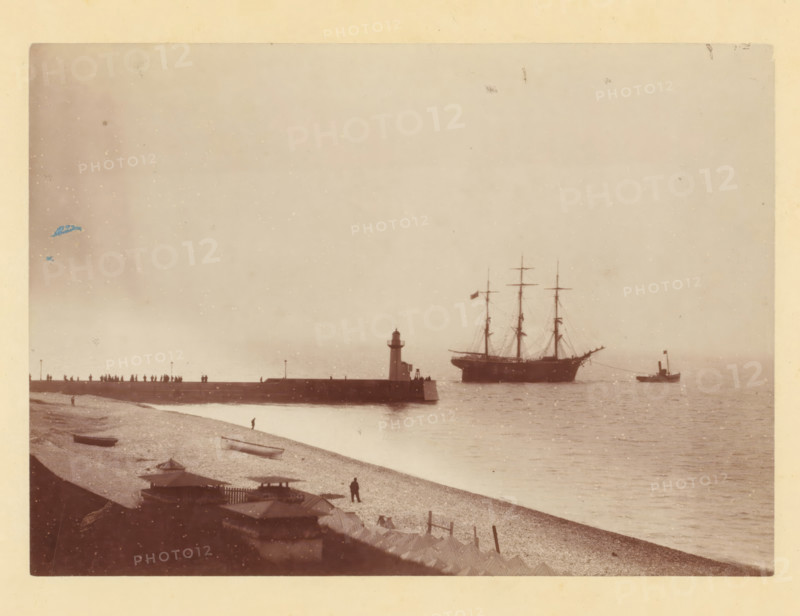 Tugboat takes sailing ship in tow at a harbor pier, photograph, Part of a French artist's album., c. 1880 - c. 1900, photographer, pasteboard, paper, albumen print, albumen print on cardboard, height 119 mm x width 166 mm, sailing-ship, sailing-boat, pier, quay, wharf, tug, pusher tug, A faded seaside scene shows a pebble beach in the foreground with small boats resting on the shore and a solitary figure standing near the water, a line of beach huts and low structures along the edge, a long stone pier extending into the water with a small lighthouse at its end and silhouettes of people standing along the pier, a large sailing ship with tall masts anchored offshore and a smaller steam-powered vessel nearby, calm water reflecting the vessels, and a pale sky above with some scattered spots and imperfections on the photograph.