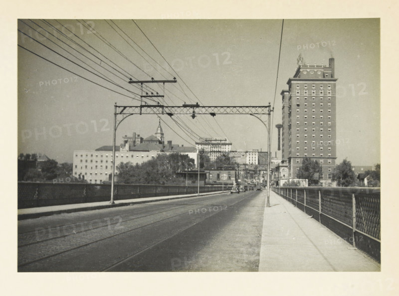 Unknown location in the United States: view in a city, Entrance of a medium-sized American city, photograph, title: 'ENTRANCE OF A MID-SIZED AMERICAN CITY', photographer: attributed to Wouter Cool, United States of America, 1936, North America, Attributed to, pasteboard, paper, gelatin silver print, developing gelatin silver print, height 157 mm x width 225 mm, height 315 mm x width 285 mm, city-view, and landscape with man-made constructions, A wide street or bridge with tram rails runs toward a distant urban area, flanked on the right by a sidewalk and a metal railing and on the left by a low barrier. Overhead metal trusses and poles span the roadway, supporting multiple parallel wires that angle across the sky. Near the horizon there are several buildings including a tall multi-story brick building with many evenly spaced windows and a decorative roofline, lower commercial and institutional buildings with varied roof shapes, and a structure with a pointed spire. A few small vehicles...