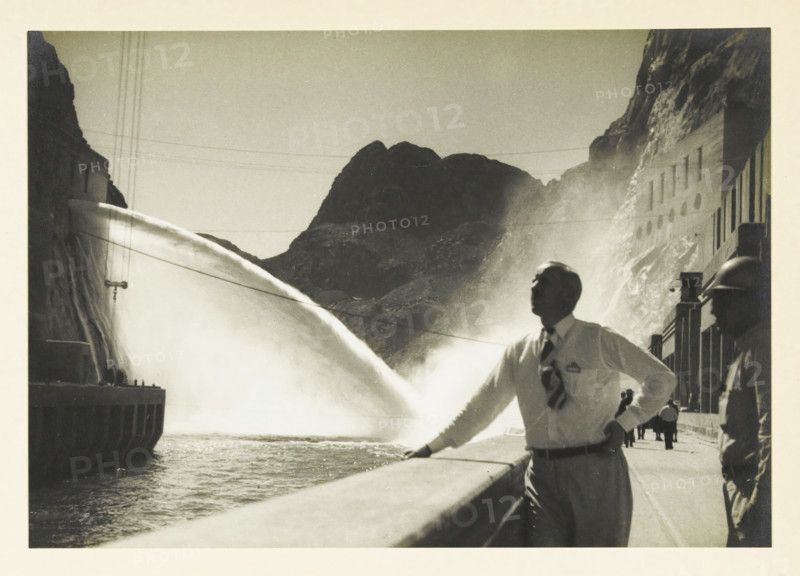 Water outlet from a power plant at an unknown location in the United States, Water outlet 60m. fall height, photograph, title: 'WATER OUTLET, 60m. FALL HEIGHT', photographer: attributed to Wouter Cool, United States of America, 1936, North America, Attributed to, pasteboard, paper, gelatin silver print, developing gelatin silver print, height 157 mm x width 225 mm, height 315 mm x width 272 mm, power station, A black and white photograph showing a large curved stream of water arcing from the left toward the center, creating mist against a backdrop of rugged dark mountains. In the foreground a man in a white shirt and patterned tie leans on a railing, his face turned upward and his hand resting on his hip, while another figure wearing a helmet stands to the right along a paved walkway with a building facade behind them. The left side shows a concrete structure with vertical elements near the water and thin cables stretching across the sky above the scene.