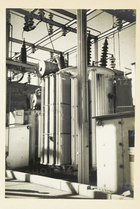 Transformer at an Electric Power Station, United States, photograph, title: '132 kV TRAFO.', photographer: attributed to Wouter Cool, United States of America, 1936, North America, Attributed to, pasteboard, paper, gelatin silver print, developing gelatin silver print, height 230 mm x width 156 mm, height 315 mm x width 330 mm, power station, A black-and-white photograph showing a section of electrical equipment with tall cylindrical and rectangular metal components, ribbed cooling fins, and large insulators mounted on a metal framework. Overhead metal beams and crossbars support suspension insulators and heavy cables that run between the structures. A vertical metal column stands in the foreground partially obscuring the equipment behind it. Shadows fall across the concrete base and the side of an enclosed cabinet with visible hinges and fasteners. The scene has a high-contrast, slightly grainy appearance.