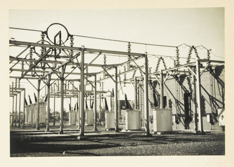 Switch installation of current-voltage transformers and isolating switches, Transformers and switches at an unknown location in the United States, photograph, title: 'SWITCH INSTALLATION OF CURRENT-VOLTAGE TRANSFORMERS, AND ISSUING SWITCHES', photographer: attributed to Wouter Cool, United States of America, 1936, North America, Attributed to, pasteboard, paper, gelatin silver print, developing gelatin silver print, height 155 mm x width 227 mm, height 315 mm x width 330 mm, power station, A wide outdoor industrial area with a grid of metal frameworks, insulators, and cylindrical tanks beneath overhead wires, with shadows cast on a gravel ground and a brick building partially visible at the right.