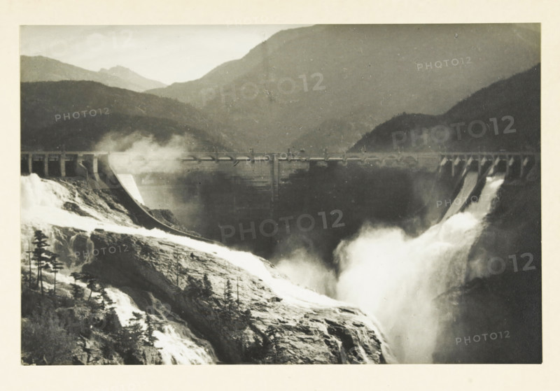 Diablo Dam and Skagit River, Washington state, United States, Skagit River project Diabolo dam, photograph, title: 'SKAGIT RIVER PROJECT, DIABOLO DAM.', photographer: attributed to Wouter Cool, United States of America, 1936, North America, Attributed to, pasteboard, paper, gelatin silver print, developing gelatin silver print, height 150 mm x width 229 mm, height 315 mm x width 330 mm, dam (in canal), A black-and-white photograph showing a large concrete dam spanning a river valley with water rushing over spillways and producing mist; rugged rocky slopes and scattered trees are visible in the foreground leading down to the turbulent water, while layered mountain ridges rise in the background behind the dam.