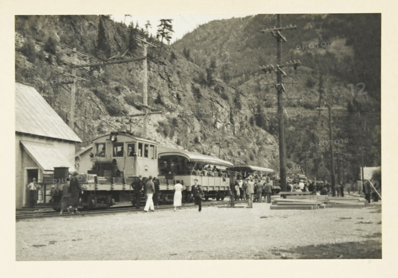 Mountain train in the valley of the Skagit River, Washington state, United States, Mountain railway through the valley of the Skagit river Seattle, photograph, title: 'MOUNTAIN STREET THROUGH THE VALLEY OF THE SKAGET RIVER SEATTLE', photographer: attributed to Wouter Cool, United States of America, 1936, North America, Attributed to, pasteboard, paper, gelatin silver print, height 152 mm x width 227 mm, height 315 mm x width 285 mm, passenger train, A black and white photograph showing a train stopped along tracks with an open-sided passenger car and a locomotive at the front, a group of people standing and milling near the train, some dressed in light clothing and some in darker coats, a building with a sloped roof to the left, telegraph poles with crossarms rising beside the tracks, wooden planks and other materials stacked near the right side of the scene, and steep tree-covered rocky hills forming the backdrop.