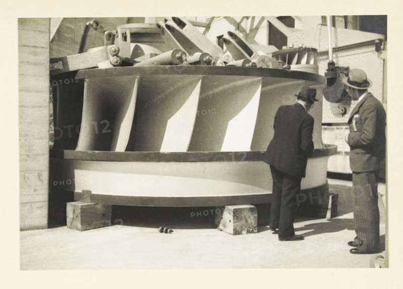 Ariel Dam, Washington state, United States: rotor of the Francis turbine, Rotor of the Francis turbine of Ariel Dam power plant, photograph, title: 'ROTOR OF THE FRANCIS TURBINE, OF ARIEL DAM POWER PLANT', photographer: attributed to Wouter Cool, United States of America, 1936, North America, Attributed to, pasteboard, paper, gelatin silver print, height 153 mm x width 224 mm, height 315 mm x width 285 mm, dam (in canal), A large circular metal structure with curved vertical blades rests on wooden blocks on a concrete floor, showing a patterned surface of alternating light and dark blade faces and a thick rim. Two men in suits and hats stand beside the structure, one facing the rim and leaning slightly forward while the other stands upright holding something near his chest. Behind the structure there are mechanical parts and structural beams with riveted plates and a cylindrical component near the upright man. Small round objects lie on the floor near the base of the structure.