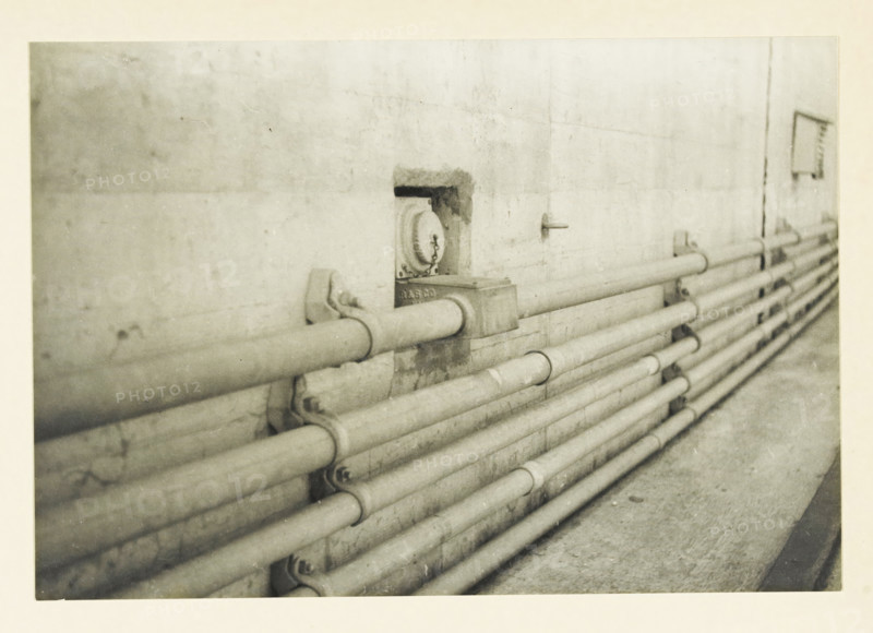 Low-voltage installation on the Ariel Dam, Ariel Dam, Washington state, United States: low-voltage installation, photograph, title: 'LOW-VOLTAGE INSTALLATION, ON THE ARIEL DAM', photographer: attributed to Wouter Cool, United States of America, 1936, North America, Attributed to, pasteboard, paper, gelatin silver print, developing gelatin silver print, height 152 mm x width 222 mm, height 315 mm x width 285 mm, dam (in canal), A grainy black and white photograph of a concrete wall with a row of horizontal metal pipes running along its length, the pipes held by brackets and clamps and appearing worn and slightly rusted, a recessed round fixture set into the wall with a chain hanging from it, a small rectangular box attached near the pipes, and a narrow ledge or walkway below the pipes.