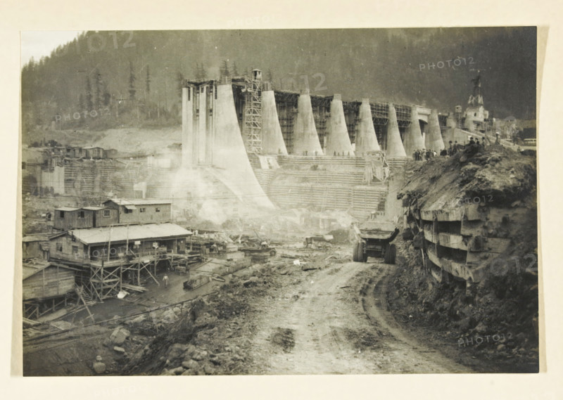 Bonnerville Dam, Portland, Oregon, United States, Portland Oregon, photograph, title: 'BONNERVILLE DAM, PORTLAND-OREGON', photographer: attributed to Wouter Cool, United States of America, 1936, North America, Attributed to, pasteboard, paper, gelatin silver print, developing gelatin silver print, height 152 mm x width 226 mm, height 315 mm x width 330 mm, dam (in canal), A wide black-and-white scene shows a large construction site with a partially built concrete structure made of tall tapered vertical supports and scaffolding, terraces and stepped forms in front of it, and a dirt road running toward the foreground. On the left there are wooden buildings on stilts with pitched roofs and external stairways, and scattered construction materials and equipment around them. A heavy dump truck sits on the dirt road, which is flanked on the right by a steep earthen embankment reinforced with horizontal wooden planks and a group of people standing along the top. In the background a forested hi...