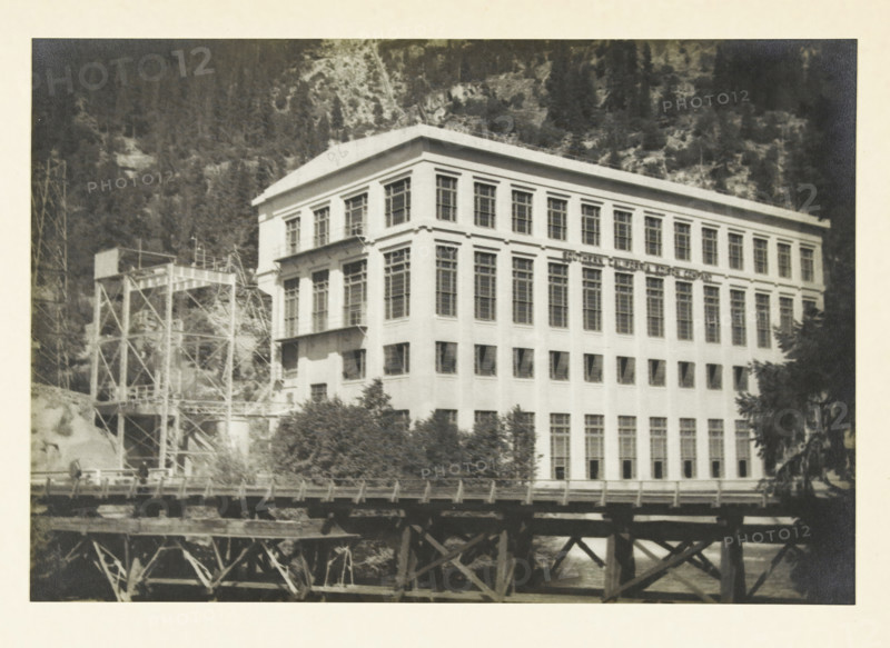 Centrale a, d Big Creek, Fresno California, Big Creek Power Plant, Fresno, California, United States, photograph, title: 'CENTRALE A, D BIG CREEK, FRESNO CALIFORNIE', photographer: attributed to Wouter Cool, United States of America, 1936, North America, Attributed to, pasteboard, paper, gelatin silver print, developing gelatin silver print, height 156 mm x width 228 mm, height 315 mm x width 285 mm, power station, A large multi-story rectangular building with many tall windows occupies the center of the image, its light-colored facade showing rows of uniformly spaced window openings and decorative cornice lines near the roof, and a flat roof. To the left of the building a metal scaffolding or industrial framework with platforms and diagonal supports rises up, partially obscured by trees and vegetation at its base. In front of the building a wooden trestle or bridge structure with visible beams and supports spans horizontally across the lower part of the image, and water can be seen be...