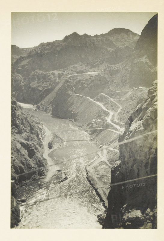 View in the Black Canyon, United States, Black Canyon, photograph, title: 'BLACK CANYON', photographer: attributed to Wouter Cool, United States of America, 1936, North America, Attributed to, pasteboard, paper, gelatin silver print, developing gelatin silver print, height 227 mm x width 153 mm, height 315 mm x width 212 mm, ravine, chasm, abyss, canyon, A tall vertical black-and-white photograph of a deep rocky canyon with steep cliffs on both sides and a winding river flowing through the center, bright reflective water visible along its length, narrow switchback roads carved into the canyon walls, layered rugged mountain ridges in the background under a pale sky, and rough textured rock faces with visible striations and ledges in the foreground.