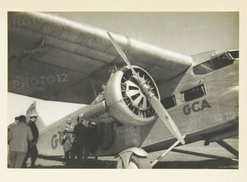 Our plane for the flight over the Grand Canyon, Aircraft before departure for a flight over the Grand Canyon, United States, photograph, title: 'OUR PLANE FOR THE, FLIGHT OVER THE GRAND CANYON', photographer: attributed to Wouter Cool, United States of America, 1936, North America, Attributed to, pasteboard, paper, gelatin silver print, height 154 mm x width 226 mm, height 315 mm x width 285 mm, aircraft, aeroplane, A black and white photograph showing a close-up of a corrugated metal airplane wing and fuselage with a radial engine and propeller mounted beneath the wing, the engine cowling and strut visible with ribbed metal texture, a portion of the cockpit window and passenger windows on the fuselage, the landing gear strut and wheel at the bottom, and a group of people standing beside the plane near the entrance, some wearing hats and coats.