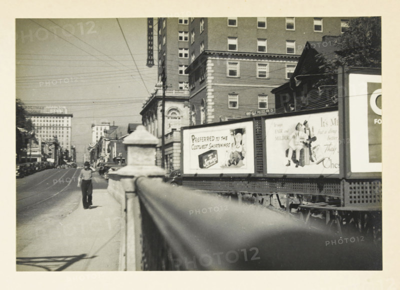 Knoxville, View in Knoxville, Tennessee, United States:, photograph, title: 'KNOCKSVILLE', photographer: attributed to Wouter Cool, United States of America, 1936, North America, Attributed to, pasteboard, paper, gelatin silver print, developing gelatin silver print, height 158 mm x width 227 mm, height 315 mm x width 285 mm, street (city(-scape) with figures, staffage), A black and white street scene showing a sidewalk with a stone railing in the foreground leading toward a distant figure walking away, a multi-story brick building and a taller building behind it along the right side, a wooden structure with a pitched roof and trees adjacent to the buildings, a row of billboards mounted on a raised platform next to the street displaying illustrated advertisements and an image of a seated person, a roadway with parked cars and streetcar tracks extending into the distance, a distant cluster of larger buildings and a hotel sign visible on the left skyline, numerous overhead utility lines ...