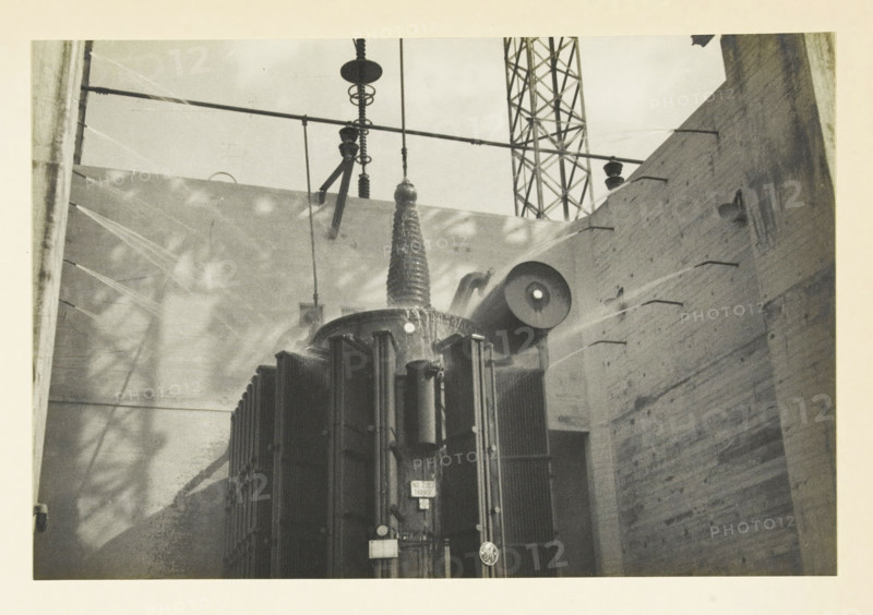 Safe Harbor Dam, Pennsylvania, United States: fire extinguishing system, Fire extinguishing system of transformers at Safe Harbor, photograph, title: 'FIRE EXTINGUISHING INSTALLATION OF, TRANSFORMERS AT SAFE HARBOR.', photographer: attributed to Wouter Cool, United States of America, 1936, North America, Attributed to, pasteboard, paper, gelatin silver print, developing gelatin silver print, height 153 mm x width 227 mm, height 315 mm x width 330 mm, dam (in canal), A large industrial transformer with vertical cooling fins and cylindrical attachments sits between two tall brick walls, with insulators and suspension hardware extending upward from its top toward horizontal crossarms; a lattice steel tower is visible in the background, and light and shadow patterns fall across the walls and transformer surfaces.
