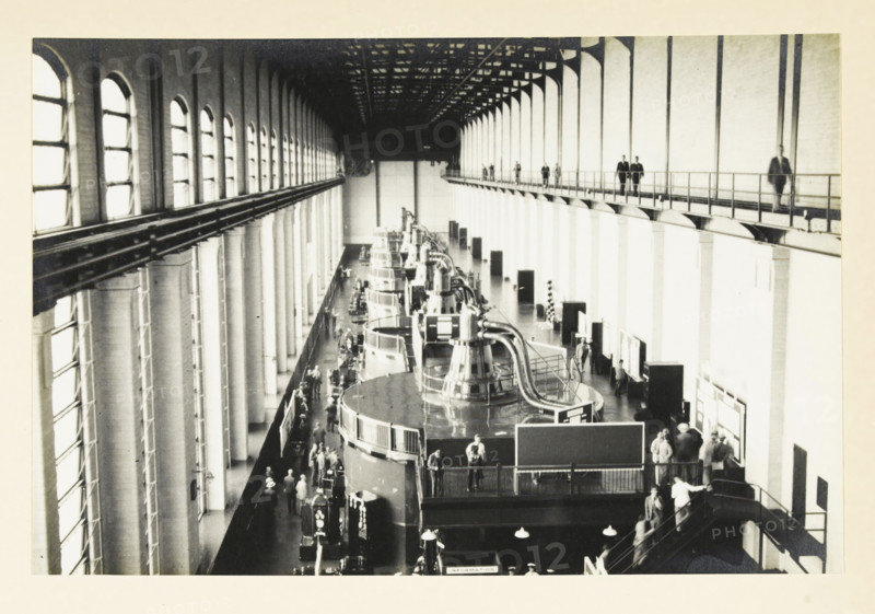 Safe Harbor Dam, Pennsylvania, United States: interior of the power station, Interior Safe Harbor power station, photograph, title: 'INTERIOR POWER PLANT, SAFE HARBOR.', photographer: attributed to Wouter Cool, United States of America, 1936, North America, Attributed to, pasteboard, paper, gelatin silver print, developing gelatin silver print, height 153 mm x width 231 mm, height 315 mm x width 285 mm, dam (in canal), A large interior space with high arched windows along the left wall and tall vertical supports along the right, a long overhead catwalk with railings and several people walking on it, a row of large cylindrical machines with curved metal piping and railings down the center floor, groups of people standing and walking around the machines and along the floor, staircases and railings leading between levels, exposed ceiling trusses and beams above, and display boards and panels positioned near the right wall.