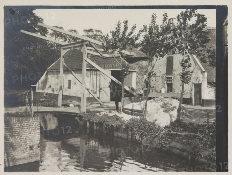 Bij Voorburg, photograph, Farm with horse and drawbridge, photographer: HC Wilkens (signed by artist), Voorburg, 1915, paper, gelatin silver print, developing gelatin silver print, height 90 mm x width 122 mm, bridge, A riverside scene showing a small bridge and a wooden drawbridge structure with long beams and supporting posts in front of buildings with tiled and thatched roofs, rough plastered walls, and varied door and window openings; a person stands near the bridge beside a horse partially obscured by the structure; young trees with leaves grow along the bank and in front of the buildings; a mound of light-colored material sits near the water edge beside a wheel and other scattered objects; the water in the foreground reflects the bridge, beams, and nearby foliage, and a brick wall lines part of the riverbank.