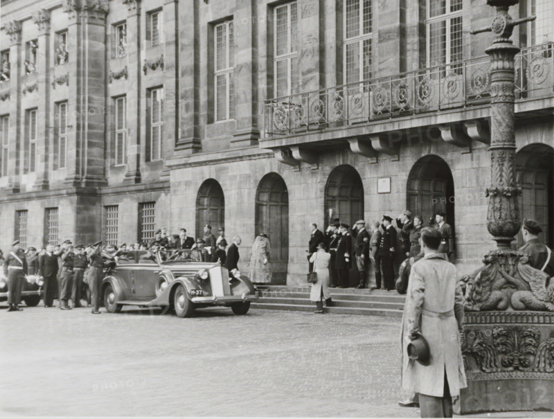 Queen Wilhelmina enters the Royal Palace in Amsterdam during her official return to Amsterdam, photograph, Part of Photo album created by Eva Pennink with photographs of Princesses Beatrix, Irene, Margriet, and Marijke from the period 1948-1951., name: 'Copyright Nederlandsch Christelijk Persfotobureau', address: 'Olympiaplein 170 Amsterdam-Z.', photographer: Nederlandsch Christelijk Persfotobureau, Dam (Amsterdam), 1945-06-28, Amsterdam, paper, gelatin silver print, height 182 mm x width 239 mm, Person, Wilhelmina of the Netherlands, queen, empress, etc. (wife of a ruler), palace, A wide stone building facade with tall windows and decorative iron balcony railings stands behind a row of arched doorways where several uniformed people and civilians are gathered on the steps; an open-top vintage car is parked in front of the building with people standing around it, and in the foreground a person in a long coat holds a hat while facing the scene near an ornate lamppost base.