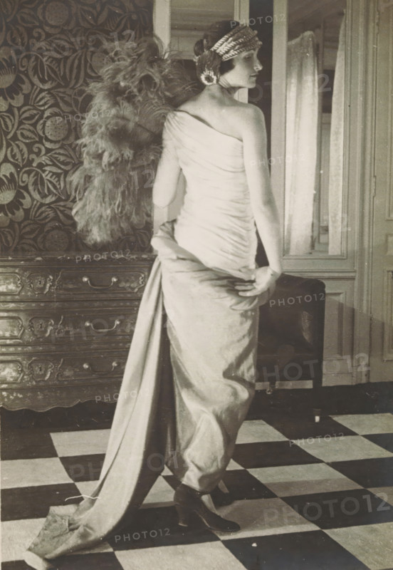 Fashion photo of a woman in a dress by Paul Poiret, Paris, photograph, inscription: '(stamp) P.Pon DE PAUL POIRET, (stamp) PAUL POIRET, 3 MARS 1923, 26, AVENUE ..., 107 FAUBOURG St. ..., (pencil and pen): 3502 Milo ?, ...', after design by Paul Poiret, 1923, photographer, paper, baryta paper, gelatin silver print, developing gelatin silver print, height 172 mm x width 122 mm, Person, model, mannequin, A black-and-white photograph of a woman standing with her back partially turned to the camera, wearing a one-shoulder draped gown that falls into a long train she holds with one hand, a decorative headband across her short hair, heeled shoes visible beneath the gown, a large feathered accessory hanging behind her, patterned wallpaper and a carved wooden chest of drawers to her side, a framed mirror or doorway and a curtain behind her, and a checkerboard floor beneath her feet.