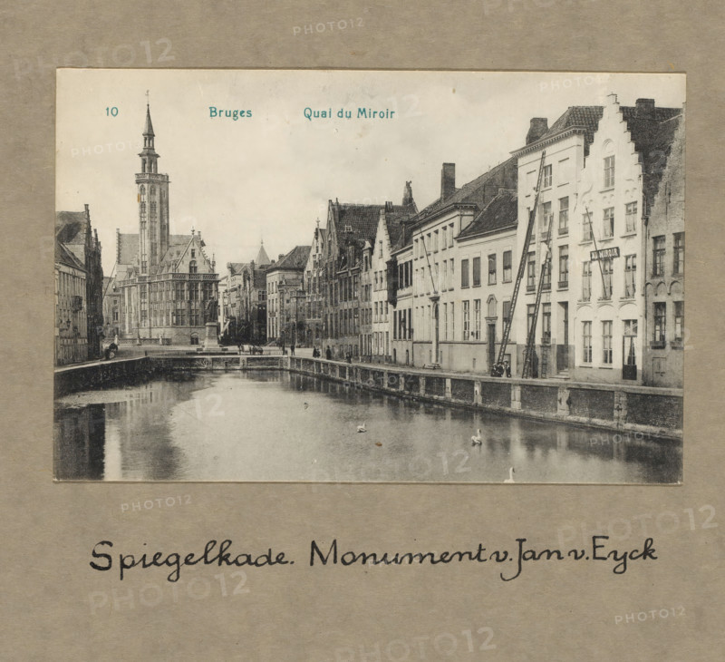 Bruges. Spiegelkade. Monument Jan van Eyck, photograph, 1860 - 1925, photographer, paper, collotype, photoprint, height 100 mm x width 150 mm, height 300 mm x width 250 mm, Person, Jan van Eyck, Bruges, A vintage black and white photograph shows a canal lined by old buildings on one side and a stone embankment on the other, with calm water reflecting parts of the scene and a few swans on the surface; the row of buildings features varied rooflines, stepped gables, many windows, and narrow facades, with ladders leaning against one façade and a small group of people near the canal edge; in the distance a tall ornate tower with a spire rises above a large ornate building with Gothic-style details, and a stone bridge and statues are visible along the canal.