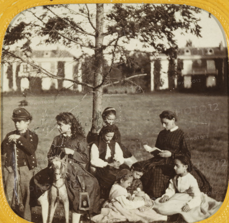 Group of people in Palace Garden, Paris, photograph, tissue stereograph, photographer: EL, France, c. 1860, paper, pasteboard, albumen print, tissue (coloured), height 86 mm x width 175 mm, height 66 mm x width 67 mm, A sepia photograph showing a group of children gathered outdoors on grass in front of a tree and a blurred building; several girls sit on a blanket, one woman or older girl holds an open book while others look down at objects in their laps, a few children wear dark dresses with white collars and one child wears a dark jacket and hat while holding a cane or staff, a small hobby horse with a visible head and reins is positioned among them, the tree trunk and branches are visible behind the group and the distant building shows arched features through foliage.