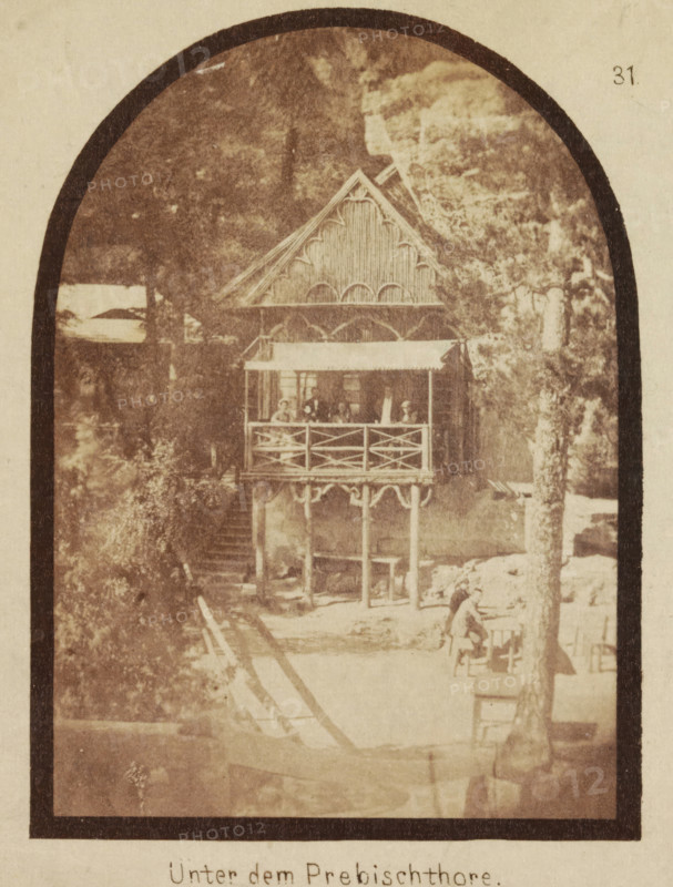 Unter dem Prebischthore, Building under the rock formation Prebischtor, stereograph, The Prebischthor (Pravcická brána) is located in the present-day Czech Republic on the border with Germany., Czech Republic, c. 1855, photographer, albumen print, salted paper print, salt print or albumen print, height 84 mm x width 171 mm, rock-formations, Pravcická brána, A sepia-toned photograph framed by a dark arched border shows a wooden stilted pavilion with decorative gables and a covered balcony railing, set against a backdrop of trees and rock formations; figures are visible on the balcony and a person stands near a tree in the foreground, with wooden posts and a walkway leading toward the structure.
