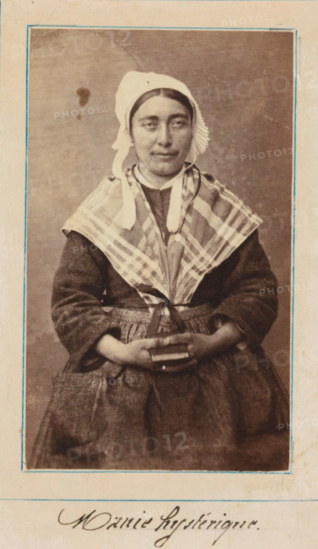 Portrait of a patient in a lunatic asylum, possibly Salpêtrière, photograph, : 'hysterical mania', c. 1860 - c. 1885, photographer, paper, albumen print, height 92 mm x width 56 mm, historical persons not known by name - BB - woman, lunatic asylum, mental home, emotional illness, derangement, madness, A sepia portrait of a seated woman wearing a light-colored cap with ties under her chin and a pleated ruffle at the side, a checked shawl draped over her shoulders, and a dark textured dress with a full skirt and gathered apron; her hands are clasped in her lap holding a small rectangular object, and she looks slightly toward the camera with a neutral expression, framed by a simple border on a light background.