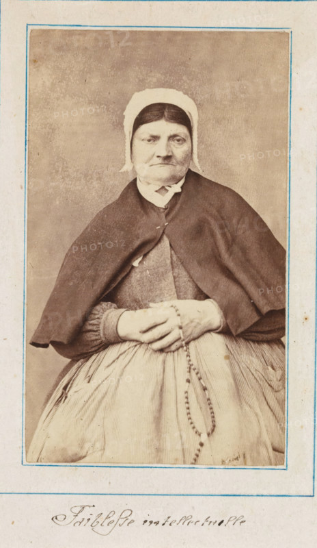 Portrait of a patient in a lunatic asylum, possibly Salpêtrière, photograph, : 'intellectual weakness', c. 1860 - c. 1885, photographer, paper, albumen print, height 92 mm x width 55 mm, historical persons not known by name - BB - woman, lunatic asylum, mental home, emotional illness, derangement, madness, A sepia-toned portrait of an older woman wearing a plain white cap and a dark shawl over a dress, seated with her hands folded in her lap holding a beaded rosary, framed by a thin blue-lined border on aged paper.