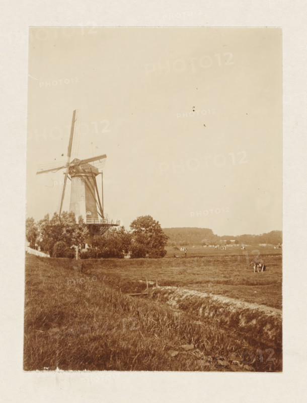 Windmill of the Schieveen polder near Overschie, photograph, Meadow with the upper windmill of the Schieveen polder near Overschie. Part of the loose-leaf photo album with 39 photos of a trip through the Netherlands, ca. 1907-1910., Netherlands, c. 1907 - c. 1910, photographer, paper, pasteboard, daylight collodion silver print, height 110 mm x width 80 mm, height 180 mm x width 235 mm, meadow, pasture, windmill in landscape, Rotterdam, Overschie, Period, 1907 - 1910, A sepia-toned photograph showing a traditional windmill with large sails on the left side, surrounded by trees and a fence at its base, an open grassy field extending from the foreground toward the right, a group of people standing in the field near the middle ground, scattered distant buildings and trees along the horizon, and an expanse of clear sky occupying the upper portion of the image.