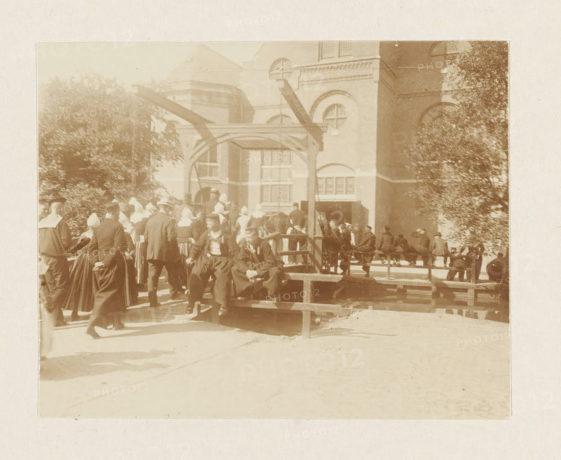 Churchgoers in Volendam, photograph, Men and women in traditional costume cross the drawbridge to the St. Vincentius Church in Volendam. Part of the loose-leaf photo album with 39 photos of a trip through the Netherlands, ca. 1907-1910., Volendam, c. 1907 - c. 1910, photographer, paper, pasteboard, daylight collodion silver print, height 80 mm x width 110 mm, height 180 mm x width 235 mm, folk costume, regional costume, church (exterior), going to church, village (city(-scape) with figures, staffage), St. Vincentius Church (Volendam), A sepia-toned outdoor scene shows a group of people gathered around a wooden platform and a simple wooden frame structure in front of a large brick building with arched windows and decorative masonry. Many figures wear long dark garments and white head coverings, while others wear dark suits and hats. Trees with dense foliage flank the scene on both sides. Some people are seated on the platform while others stand or walk nearby. The ground in front of the...