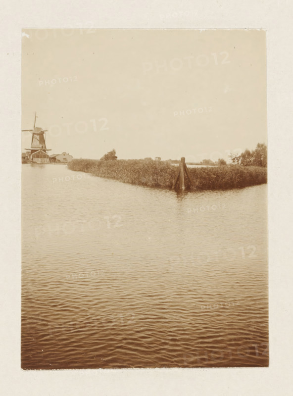 Windmill by the water (former title), Sawmill De Vriendschap in Monnickendam, photograph, View over the water with a windmill and a dolphin. Part of the loose-leaf photo album with 39 photos of a trip through the Netherlands, ca. 1907-1910., Netherlands, c. 1907 - c. 1910, photographer, paper, pasteboard, daylight collodion silver print, height 110 mm x width 80 mm, height 180 mm x width 235 mm, windmill in landscape, Monnickendam, A sepia-toned photograph showing a calm body of water in the foreground with gentle ripples, a narrow grassy peninsula extending into the water toward the middle distance, a wooden pile structure at the tip of the peninsula, reeds and low vegetation along the shoreline, a windmill and a few low buildings visible on the far left shoreline, and an open pale sky occupying the upper portion of the image.