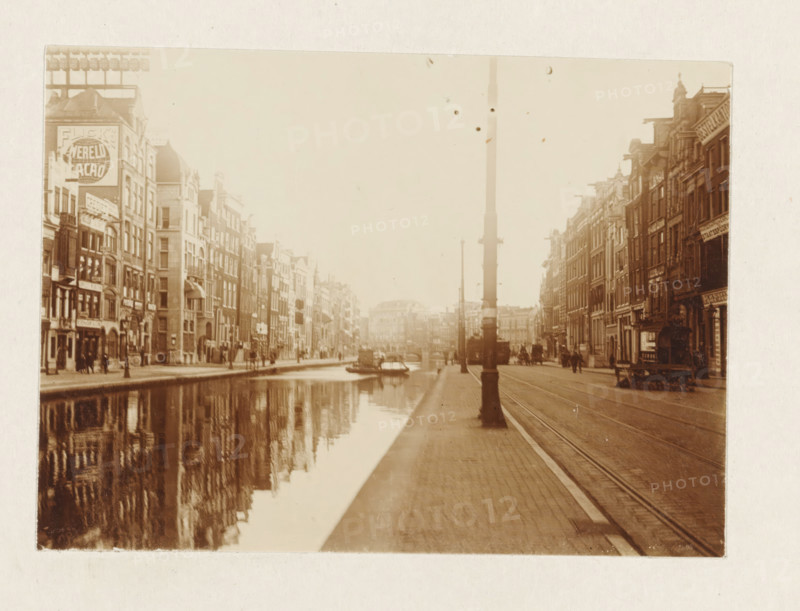 Rokin in Amsterdam, photograph, View over the Rokin in Amsterdam. Part of the loose-leaf photo album with 39 photos of a trip through the Netherlands, ca. 1907-1910., Amsterdam, c. 1907 - c. 1910, photographer, paper, pasteboard, daylight collodion silver print, height 80 mm x width 110 mm, height 180 mm x width 235 mm, canals, waters (in city) (city(-scape) with figures, staffage), street (city(-scape) with figures, Rokin, A sepia-toned street scene shows a canal running alongside rows of multi-story buildings with narrow facades and varied rooflines, their reflections visible in the water; a paved sidewalk and tram tracks run parallel to the canal with a tall streetlamp in the foreground; small boats are moored on the canal and a horse-drawn carriage is visible on the street; people stand and walk along the sidewalks and storefronts line the buildings on both sides.