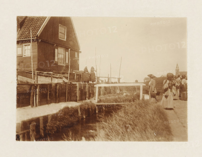 Marken, photograph, Tourists view houses in Marken; two women in traditional costume stand in front of the house. Part of the loose-leaf photo album with 39 photos of a trip through the Netherlands, ca. 1907-1910., c. 1907 - c. 1910, photographer, paper, pasteboard, daylight collodion silver print, height 80 mm x width 110 mm, height 180 mm x width 235 mm, folk costume, regional costume, A sepia photograph shows a waterside scene with a wooden house on stilts at the left edge, featuring a tiled roof and multi-paned windows, and a wooden railing and platform along the water. Two figures in dark clothing stand on the platform near tall thin poles. In the foreground a narrow water channel runs alongside grassy banks and a simple wooden bridge spans the channel. On the right several people wearing long coats and hats stand or walk along a dirt path, and a distant building with a tall pointed tower is visible on the horizon.