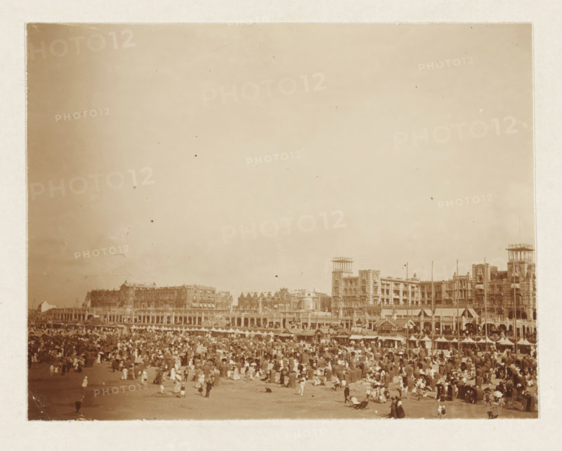 Beach at Scheveningen, photograph, Beach at Scheveningen. Part of the loose-leaf photo album with 39 photos of a trip through the Netherlands, ca. 1907-1910., Scheveningen, c. 1907 - c. 1910, photographer, paper, pasteboard, daylight collodion silver print, height 80 mm x width 110 mm, height 180 mm x width 235 mm, beach (as place of recreation), A sepia-toned photograph showing a crowded beach scene with many people scattered across the sand, many wearing long dresses and hats, some sitting or standing and a few walking; behind the beach there is a long row of multi-story buildings with ornate facades and arched colonnades, including taller structures with rooftop frameworks and towers, and a line of small beach huts or tents in front of the buildings; the sky above is mostly clear and occupies a large portion of the image.