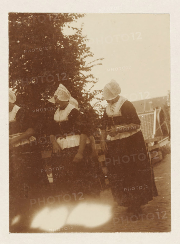 Volendam, photograph, Women in traditional costume in Volendam. Part of the loose-leaf photo album with 39 photos of a trip through the Netherlands, ca. 1907-1910., c. 1907 - c. 1910, photographer, paper, pasteboard, daylight collodion silver print, height 110 mm x width 80 mm, height 180 mm x width 235 mm, folk costume, regional costume, Three women stand side by side wearing dark dresses with light-colored aprons and intricately patterned caps covering their hair and part of their faces, one woman holds a shallow bowl against her midsection while another's hand rests near her waist, behind them a large leafy tree fills the left background and a slatted roof structure is visible on the right, the photo has a sepia tone and shows worn edges and a bright overexposed area near the bottom.