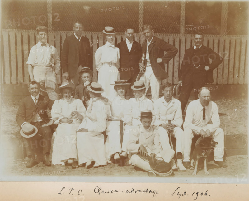 Group at tennis club LTC Quick-Advantage, Gerard Aalbersberg seated on the left, photograph, photographer: circle of Gerard Aalbersberg (us), The Hague, 1906-09, paper, daylight collodion silver print, height 130 mm x width 179 mm, A sepia photograph of a group of people posed outdoors in front of a wooden fence and wire mesh, arranged in three rows with some seated on the ground and others standing behind; men and women wear early twentieth-century clothing including light-colored dresses and blouses, suits, jackets, and straw boater hats, while one man holds a racket and another leans on a racquet, a seated man in the front holds a case, and a dog sits beside a man at the lower right.