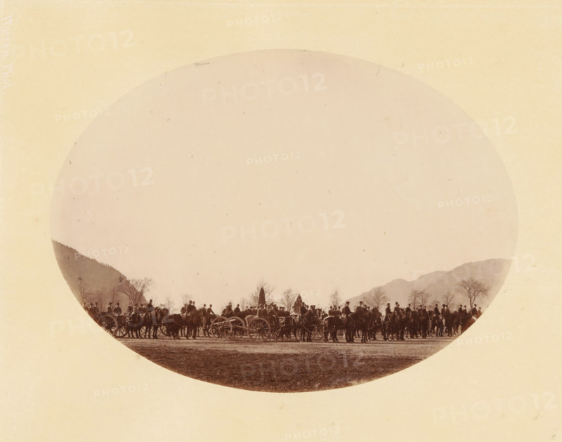 Cadets, West Point (original title), Group of Cadets of West Point, photograph, inscription: 'West Point, 1867, 225, ST1140', inscription: 'Warren Phot.', photographer: George Kendall Warren, North America, pasteboard, paper, albumen print, height 145 mm x width 198 mm, height 253 mm x width 330 mm, military instruction, American Civil War, A large circular sepia photograph centered on a pale rectangular background shows a long line of people and horses with carriages along a flat open area, with silhouettes of leafless trees and low mountains visible behind them; the scene is framed by the round edge of the photograph and the paper shows a textured, aged appearance.