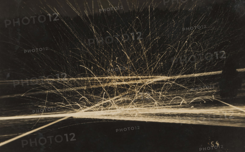 Barrage with machine gun(s) at evening or night, photograph, number: '54', name: 'PHOTO WIDMER, WALLENSTADT', inscription: 'Feu de barrage, 6 mitrailleuses, fM, ... illegible word', photographer: E. Widmer, c. 1930, paper, gelatin silver print, developing gelatin silver print, height 88 mm x width 139 mm, Dark background with many bright, thin streaks and arcs radiating upward and outward from a brighter horizontal band near the center, forming tangled, overlapping lines and curved trails that vary in length and curvature, with some straight horizontal lines crossing the scene and numerous short, arching marks concentrated near the lower center, all rendered in a pale, warm tone against the dark field.