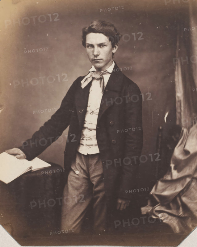 Portrait of an unknown standing young man with hand on book, photograph, photographer: attributed to Franz Hanfstaengl, Germany, c. 1855 - c. 1860, Attributed to, paper, salted paper print, salt print, height 222 mm x width 154 mm, anonymous historical person depicted, A sepia-toned portrait of a young person standing and facing the camera, wearing a dark long coat over a light-colored waistcoat and a tied neck scarf; one hand rests on an open book placed on a draped surface while the other hand hangs by the side, and folded drapery is visible to the right.