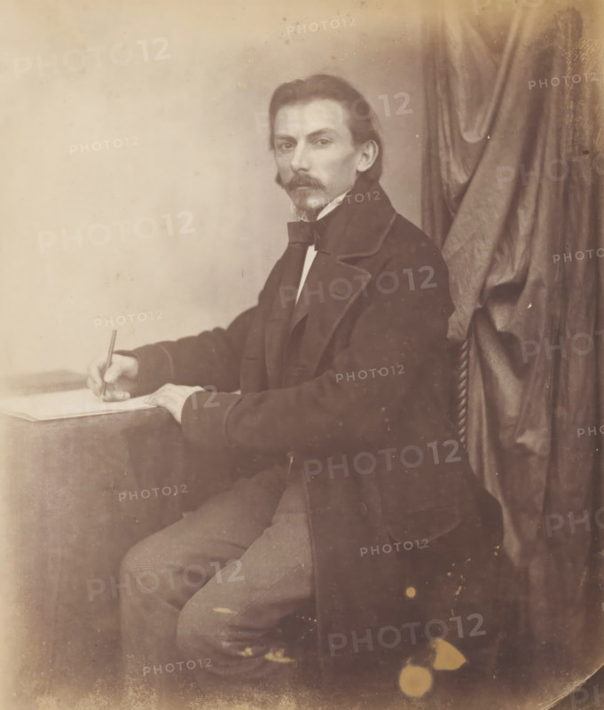 Portrait of Marggraf, architect, drawing with pencil in his hand, photograph, inscription: 'Marggraf Architekt', photographer: attributed to Franz Hanfstaengl, Germany, c. 1855 - c. 1860, Attributed to, paper, salted paper print, salt print, height 198 mm x width 163 mm, height 236 mm x width 294 mm, A seated man with a mustache and small goatee looks toward the camera while holding a pen over a sheet on a table, wearing a dark coat with wide lapels and a bow tie, with a curtain draped behind him and a plain light background on the other side, his visible arm resting on the table and his legs partially shown beneath the tablecloth.
