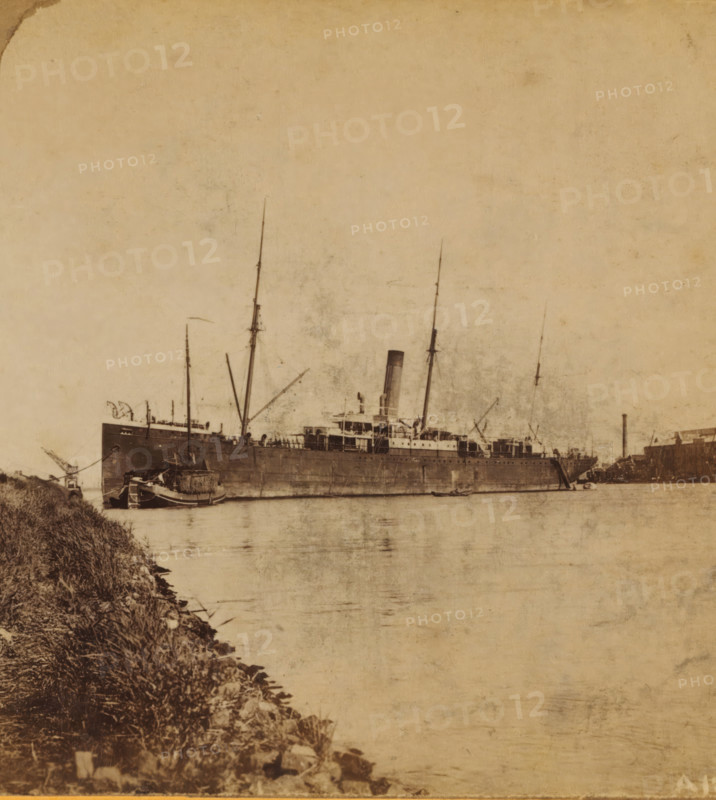 A Mailboat, View of a mailboat at the Handelskade in Amsterdam, Views and landscapes in Europe, Africa and Asia, Geographical Sculpture Gallery, collected by MJ Broekhuijsen, Headmaster of a school in Amsterdam, stereograph, Numbered A12 in the lower right corner of the photo., annotation: 'The 'Burgemeester Den Tex' of the Maatschappij Nederland at the breakwater, at the Handelskade in Amsterdam. In the foreground on the left: a hay barge.', Amsterdam, publisher: Merkelbach & Co., compiler (person in literature): Martien Johan Broekhuijsen, c. 1905, photographer, compiler (person in literature), pasteboard, paper, albumen print, height 87 mm x width 176 mm, Part of a group of 52 stereophotos used in primary education as a supplement to school charts. The entire series consisted of 274 items in 10 categories (A to K)., steamship, motorship, A large vintage steamship sits low in calm water with tall masts and a central funnel rising above a deck crowded with rigging and structures, a sm...