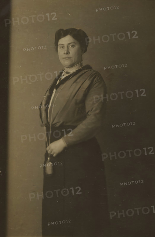 Portrait of Aagje Nieuwmeijer, quick photo, Agatha Jacoba ('Aagje') Nieuwmeijer, profession unknown, wife of Jacobus Johannes Griss, teacher of Dutch Language and Literature at a secondary school in Rotterdam, lived in Rotterdam at the time of the photo., address: 'Kalverstraat 155 E, 178', photographer: American Automatic Fotografie, Amsterdam, 1914, paper, baryta paper, gelatin silver print, developing gelatin silver print, height 86 mm x width 52 mm, Person, Aagje Nieuwmeijer, Kalverstraat, A sepia-toned portrait of a woman standing against a plain backdrop, shown from head to below the waist, with dark hair styled and parted, a composed facial expression, wearing a high-collared blouse and a long-sleeved jacket with a darker trim, a long dark skirt, and holding a small cylindrical object suspended from a cord in one hand at waist level.