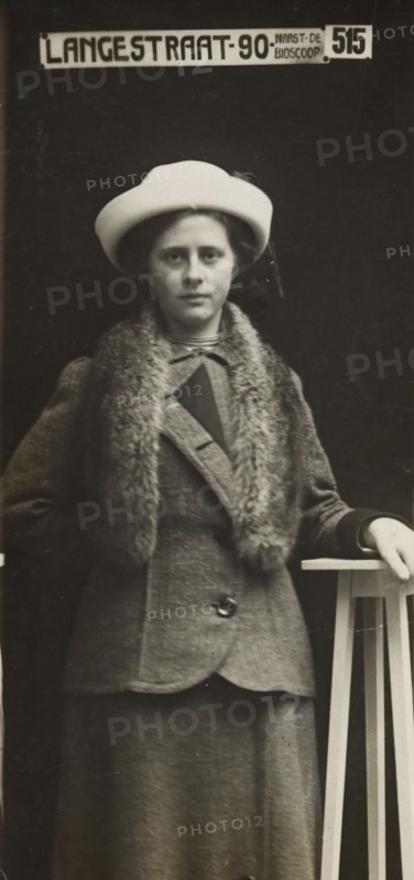 Rapid photographs, snelfoto, photographer: American Tip Top Foto Company, Alkmaar, before 1913-07-08 - after 1917-07, paper, gelatin silver print, developing gelatin silver print, height 83 mm x width 41 mm, A black and white portrait of a woman standing against a dark backdrop, wearing a light-colored hat and a coat with a fur collar, her face visible and expression neutral, one hand resting on a tall wooden stand with vertical slats, the coat showing a button and textured fabric and a skirt extending below, the image framed in a tall narrow format.